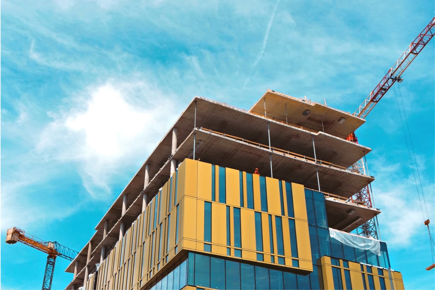 Modern building under construction with yellow and glass façade, construction cranes visible against bright blue sky.