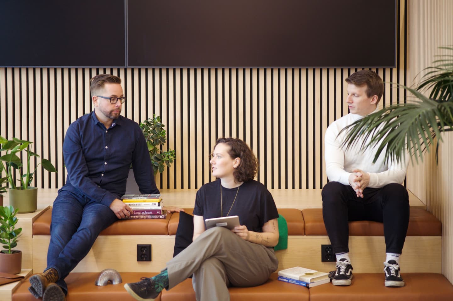 Three colleagues in conversation on a tan leather bench with wooden slat wall background and potted plants nearby.