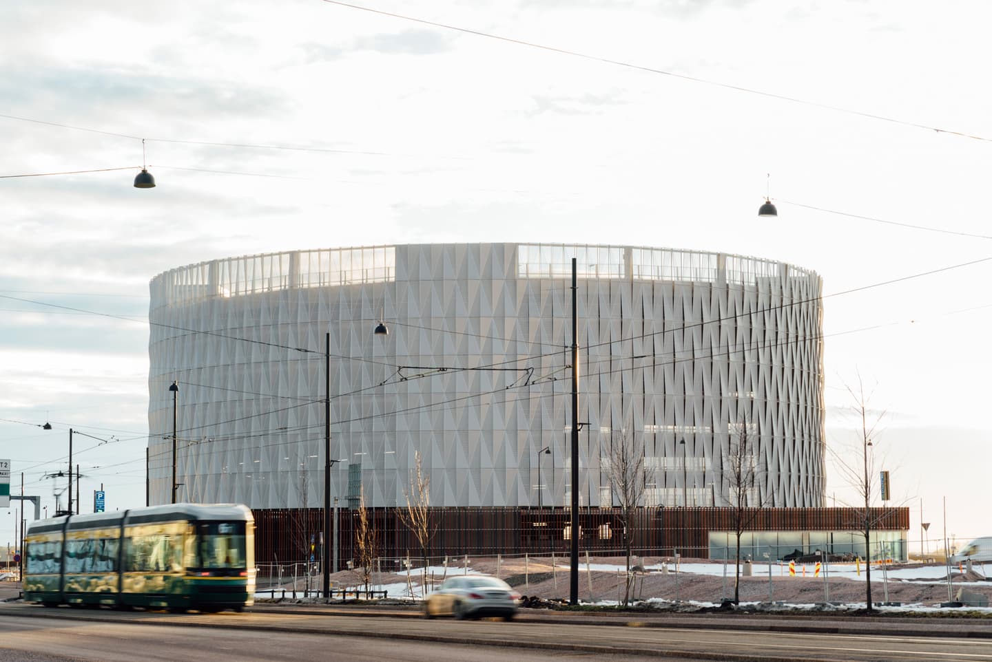 Modern circular building with geometric white façade, tram passing in foreground under cloudy sky.