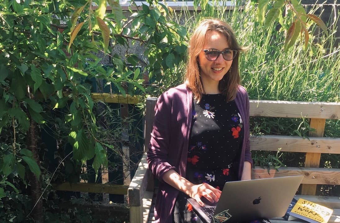 Person in sunglasses working on laptop outdoors in a lush garden setting, wearing purple cardigan over floral top.