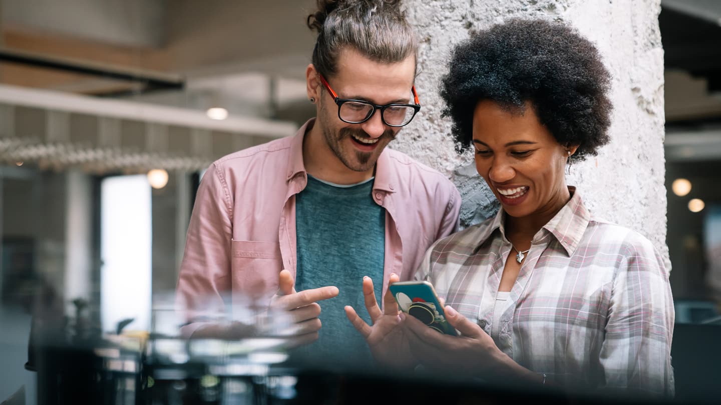 Two smiling colleagues looking at a mobile phone together in a modern office space with blurred background.