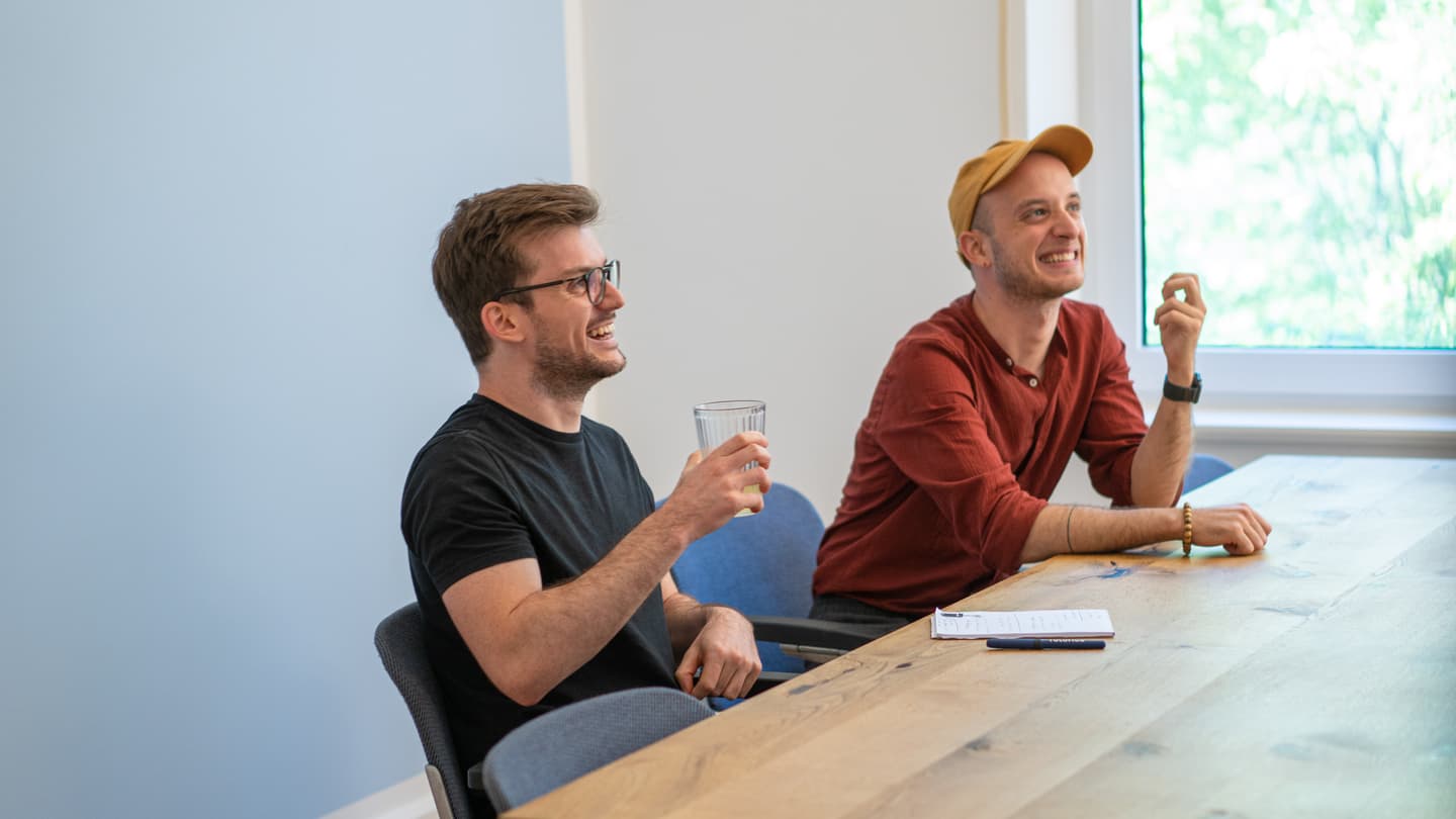 Two smiling men sitting at wooden table in bright office, one in black t-shirt holding drink, other wearing orange cap.