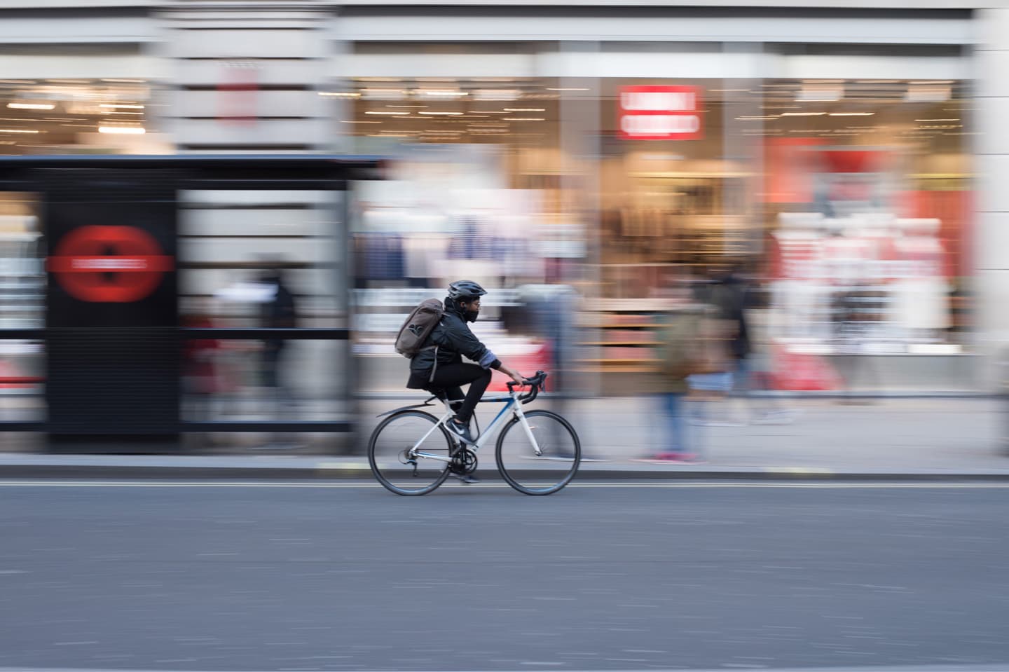 Cyclist with backpack riding a white bicycle past blurred storefronts, captured with motion effect in an urban setting.