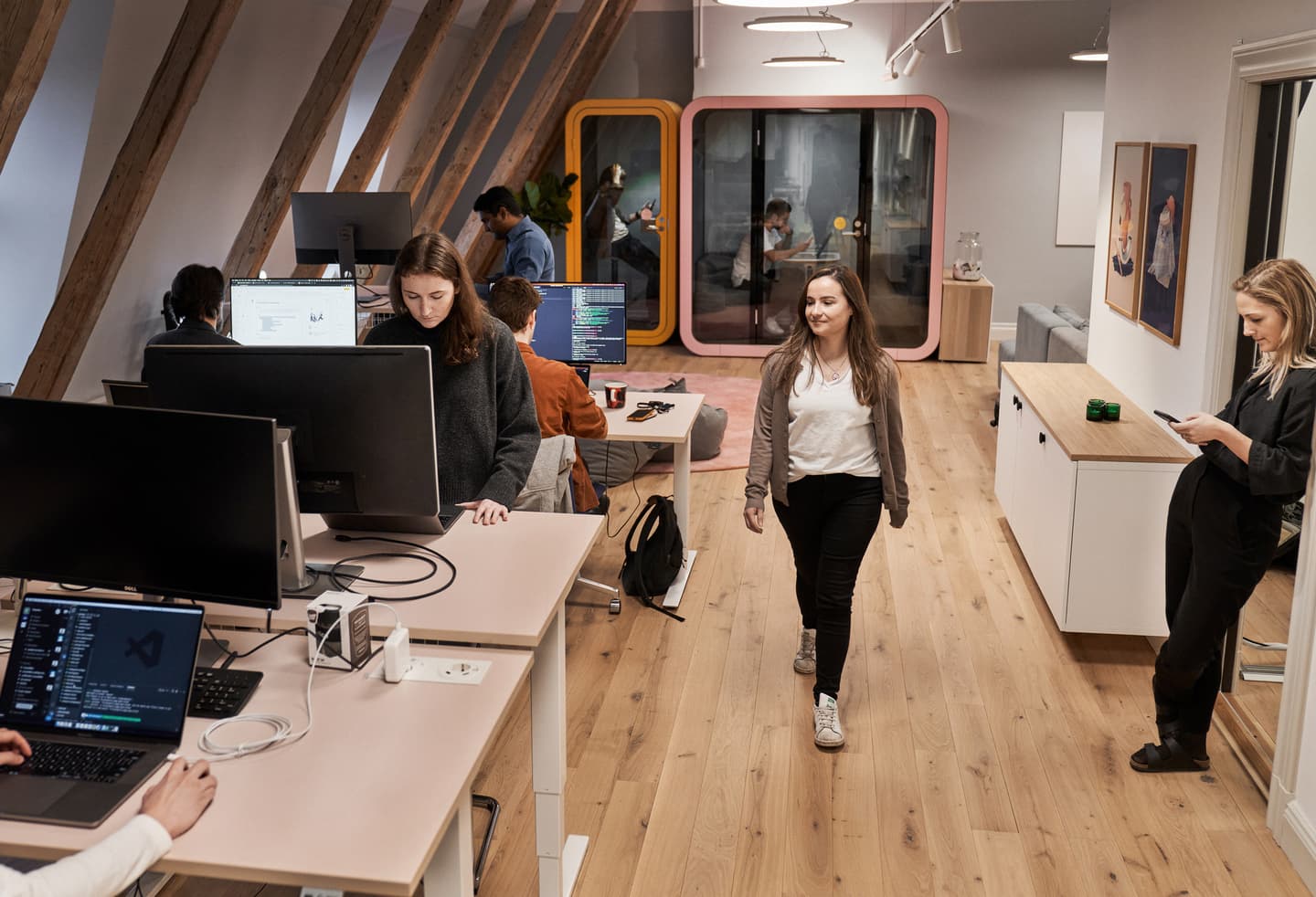 Modern office with wooden beams, people working at computers, colourful phone booths, and wooden flooring.