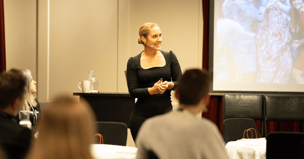Speaker in black dress with headset microphone presenting to audience with projection screen visible in background.