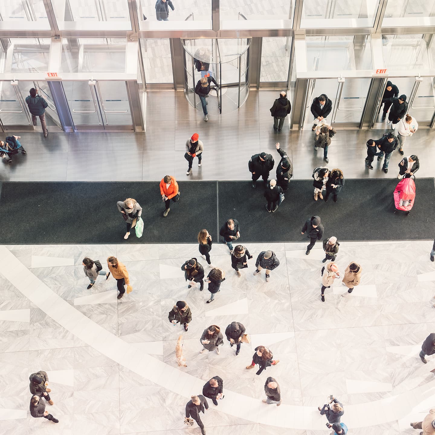 Aerial view of people walking through a modern building lobby with marble floors, glass revolving doors and exit signs.